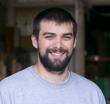 Smiling young man with a beard, wearing a gray shirt, stands in a workshop with blurred tools in the background.