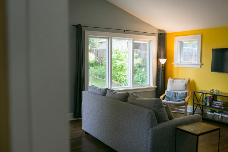 Cozy living room featuring a gray sofa, yellow accent wall, large windows, and a wooden chair beside a lamp.