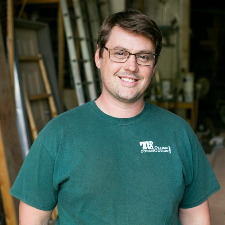 Smiling man in a green t-shirt stands in a workshop, with tools and equipment visible in the background.