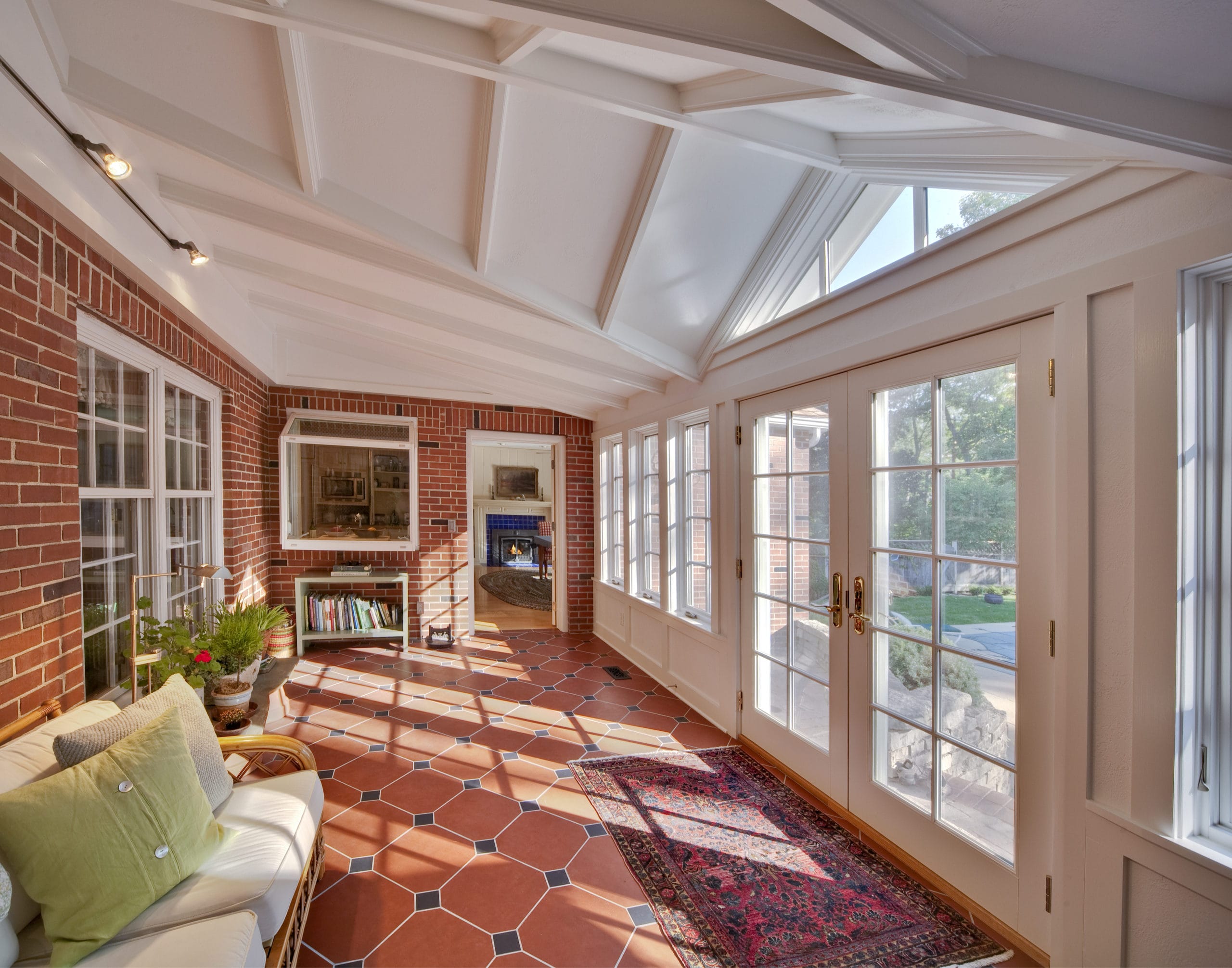 Bright sunroom with brick walls, a cozy sofa, and large windows, featuring a patterned rug and a view of the outdoors.