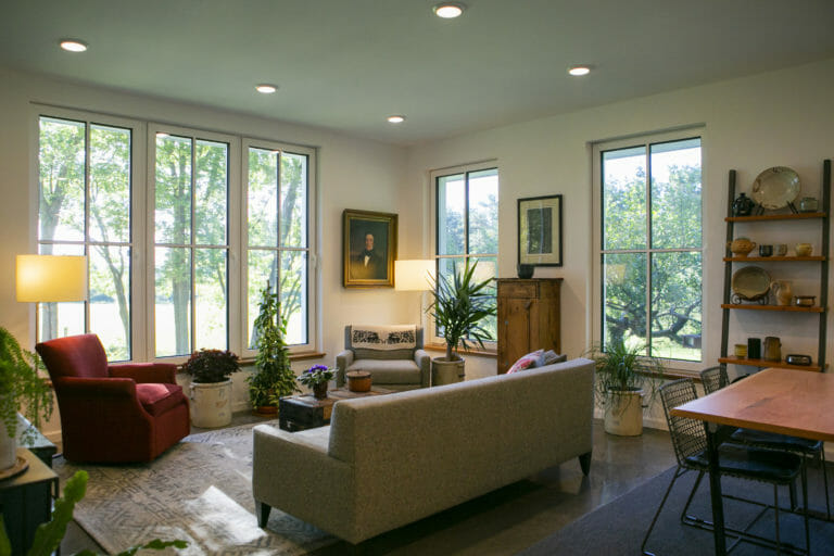 Cozy living room with large windows, plants, a red armchair, and a wooden dining table, illuminated by soft lighting.