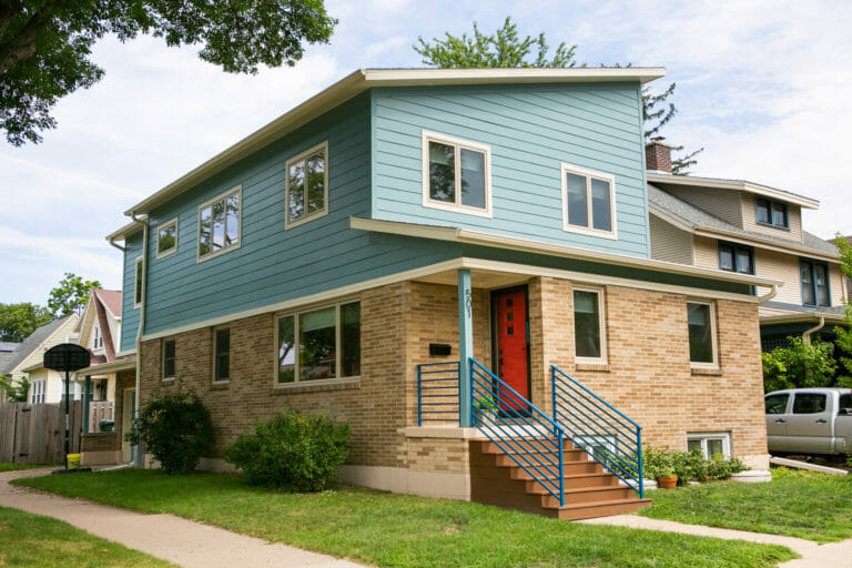 Modern two-story house with blue siding, red front door, and a landscaped yard, surrounded by neighboring homes.