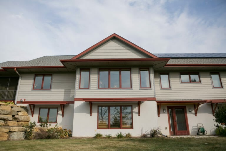 Modern two-story house with a gray exterior, red trim, large windows, and solar panels on the roof, surrounded by greenery.