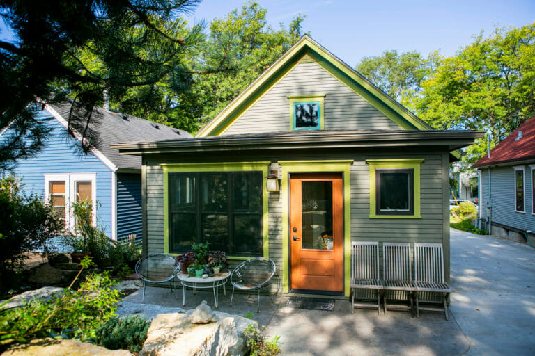 Charming green-trimmed house with a wooden door, surrounded by greenery and outdoor seating on a sunny day.