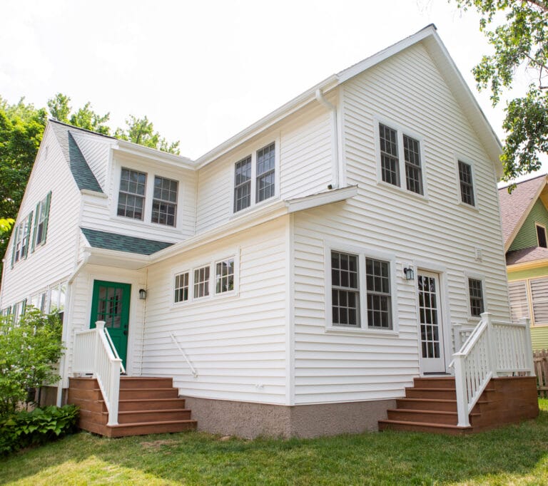 Two-story white house with green accents, featuring a porch, steps, and multiple windows surrounded by grass and trees.