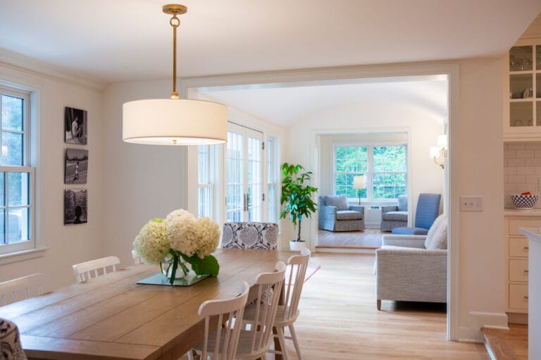 Bright dining area with a wooden table, white chairs, and a floral centerpiece, leading to a cozy living room.