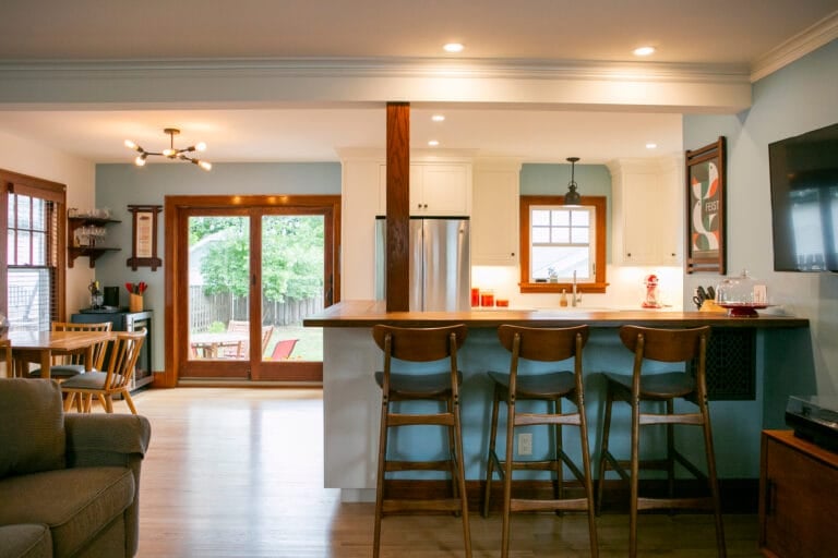 Bright, modern kitchen with wooden bar stools, open to a cozy dining area and a view of a green backyard through sliding doors.