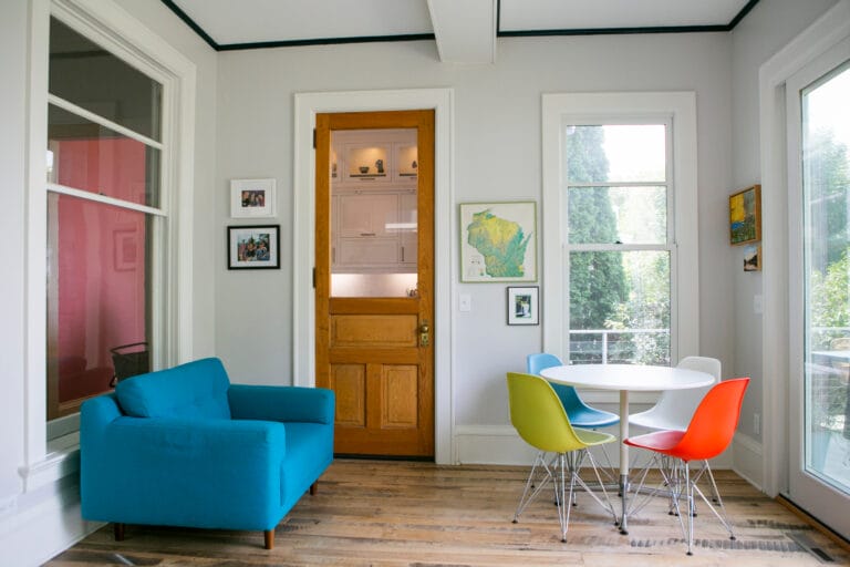 Bright dining area featuring a turquoise couch, colorful chairs, and a wooden door leading to a kitchen.