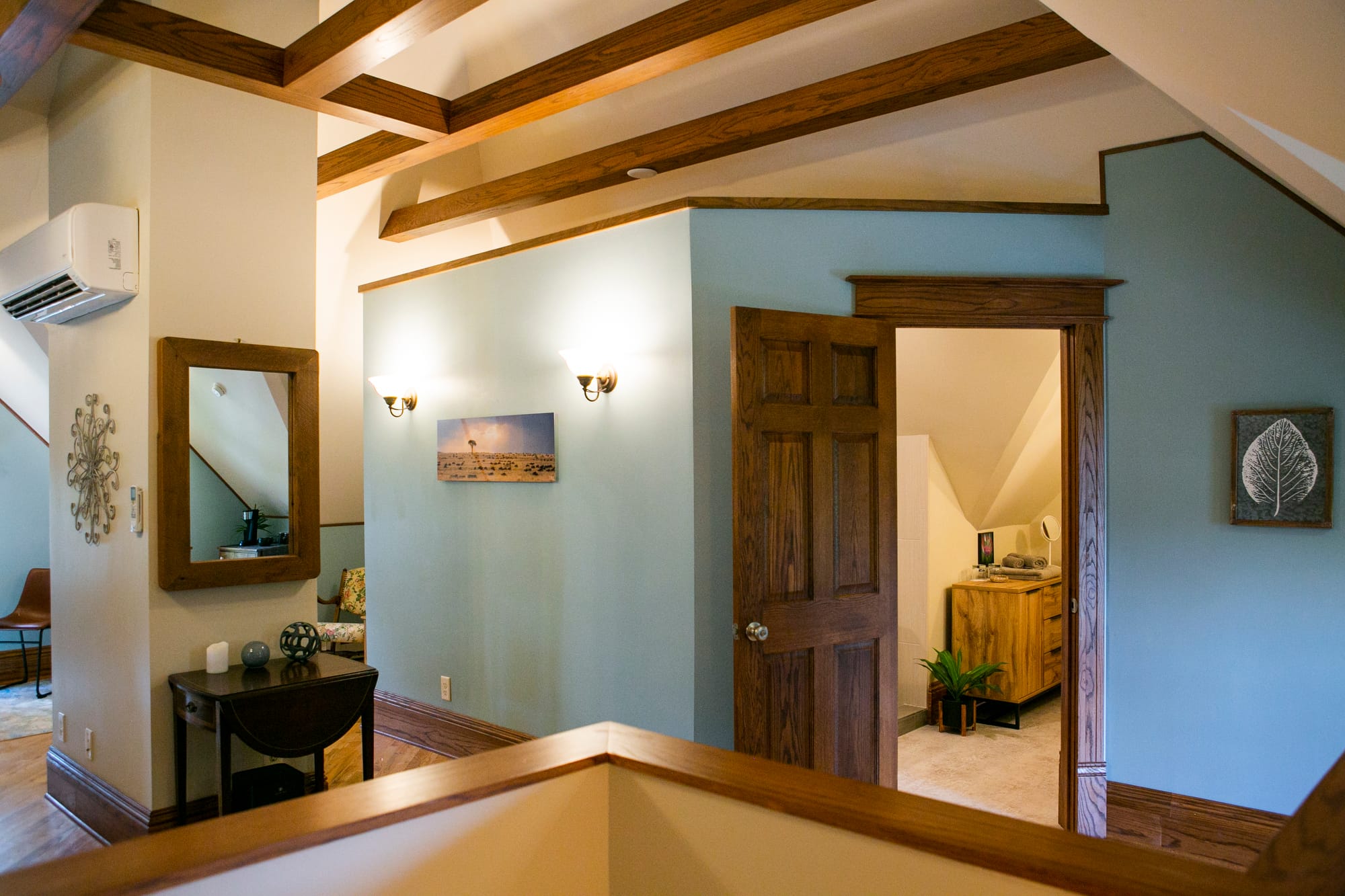 Cozy interior with wooden beams, a mirror, and a glimpse of a room beyond a wooden door, featuring soft lighting.