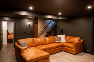 Cozy basement lounge featuring a large brown leather sectional, soft lighting, and a staircase leading to an upper level.