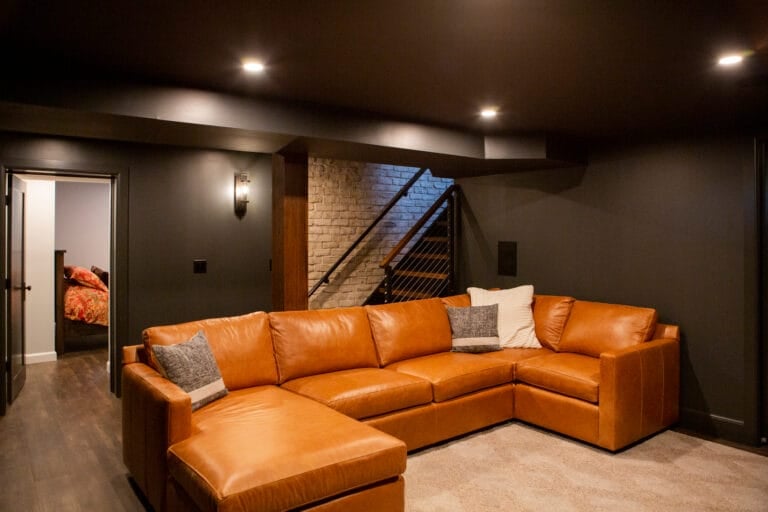 Cozy basement lounge featuring a large brown leather sectional, soft lighting, and a staircase leading to an upper level.