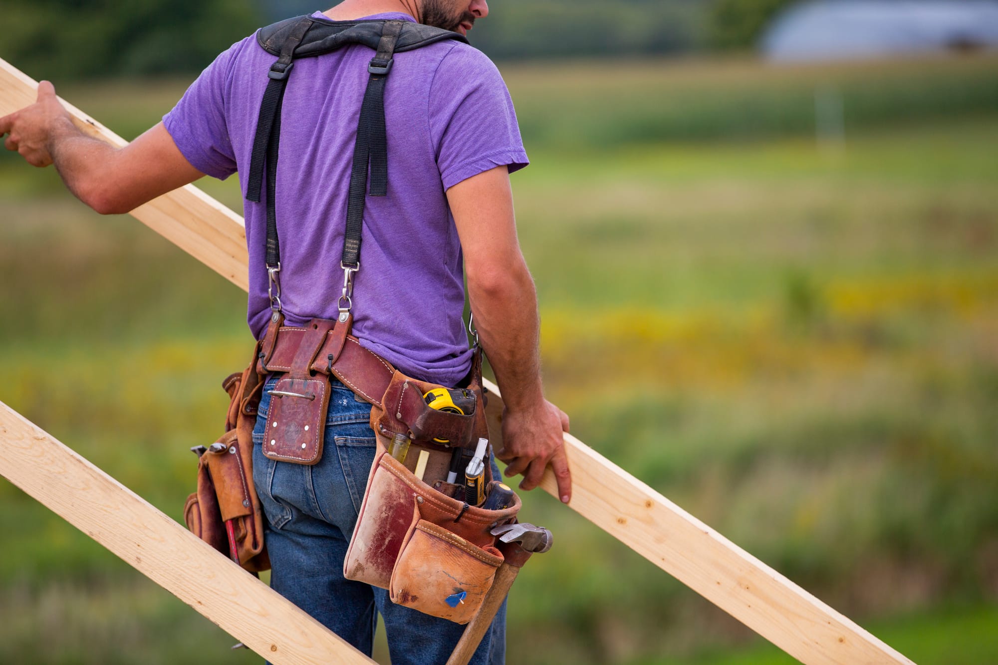 A man in a purple shirt carries a wooden beam, wearing a tool belt filled with various tools against a blurred green background.