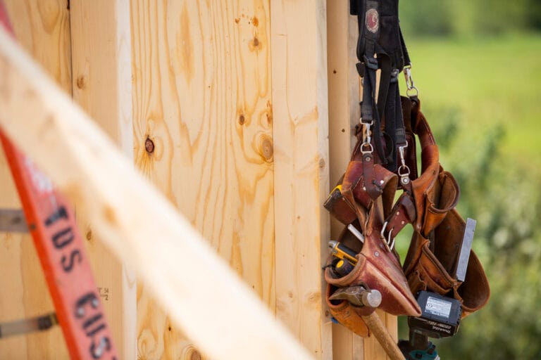 Leather tool belts hang on a wooden wall, with a ladder and green landscape blurred in the background.