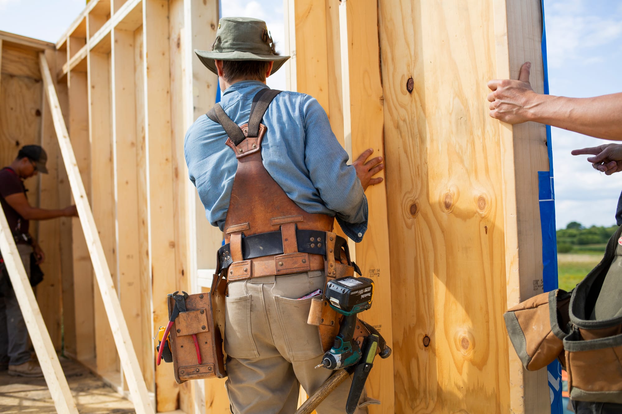 A carpenter in a blue shirt and leather tool belt adjusts a wooden wall frame, with another worker visible in the background.