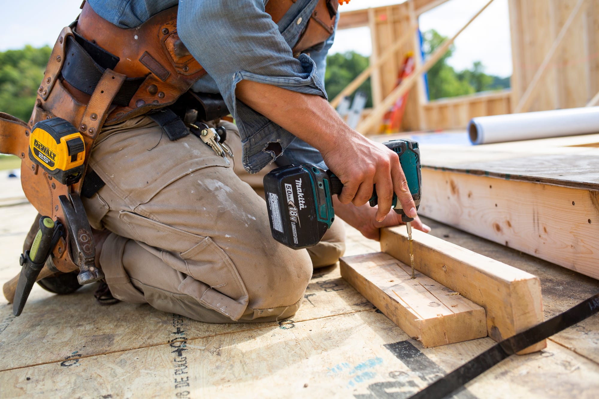 A construction worker kneels on a wooden floor, using a power drill to secure wooden pieces together.