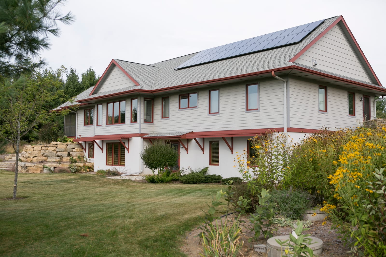Modern two-story house with solar panels, surrounded by greenery and colorful flowers in a landscaped yard.