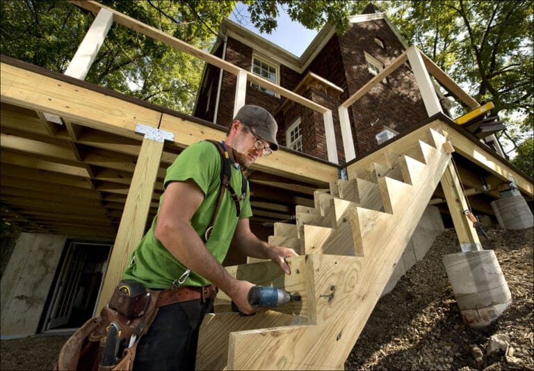 A construction worker in a green shirt installs wooden stairs on a deck under a house, surrounded by trees.