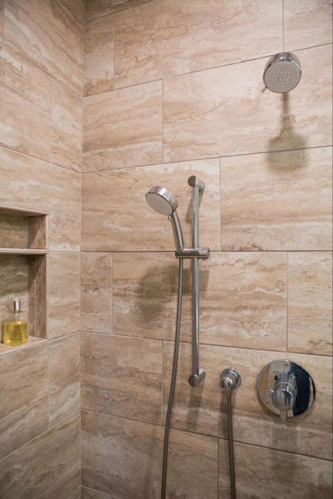 Modern shower with sleek chrome fixtures, beige stone tiles, and a built-in shelf holding a bottle of shower oil.