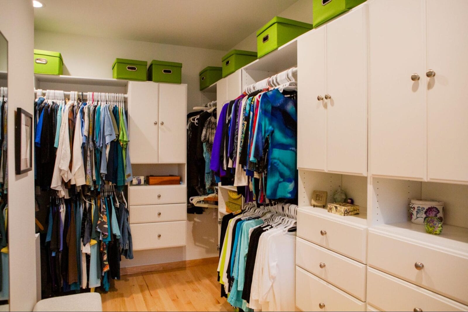 A well-organized closet featuring colorful clothing on hangers, white cabinets, and green storage boxes on top shelves.