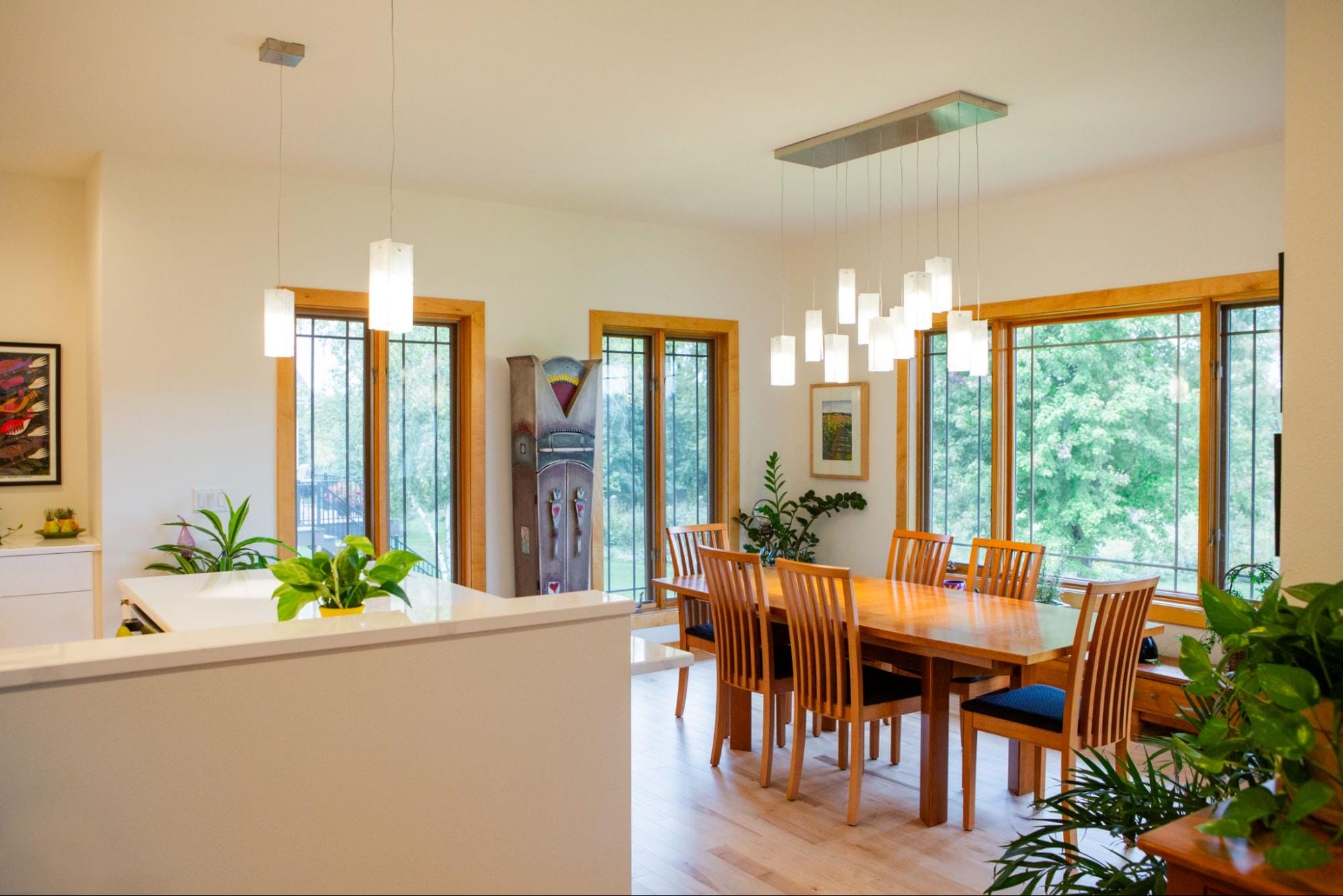 Bright dining area featuring a wooden table, stylish chairs, and large windows with greenery outside. Modern pendant lighting above.