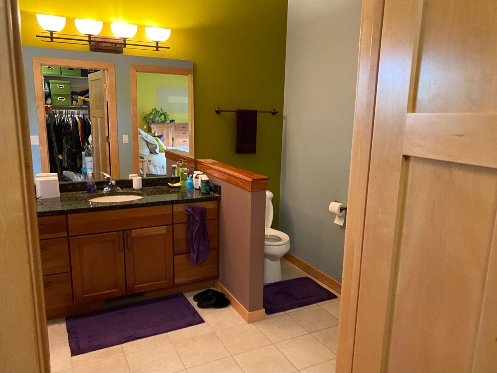 Bright bathroom featuring a wooden vanity, green accent wall, and a mirror reflecting a closet with colorful clothing.