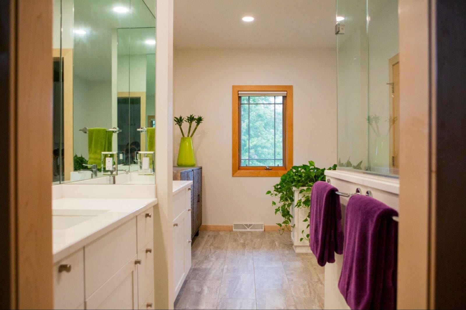 Spacious bathroom with a double vanity, green plants, and a window, featuring purple towels hanging by the sink.