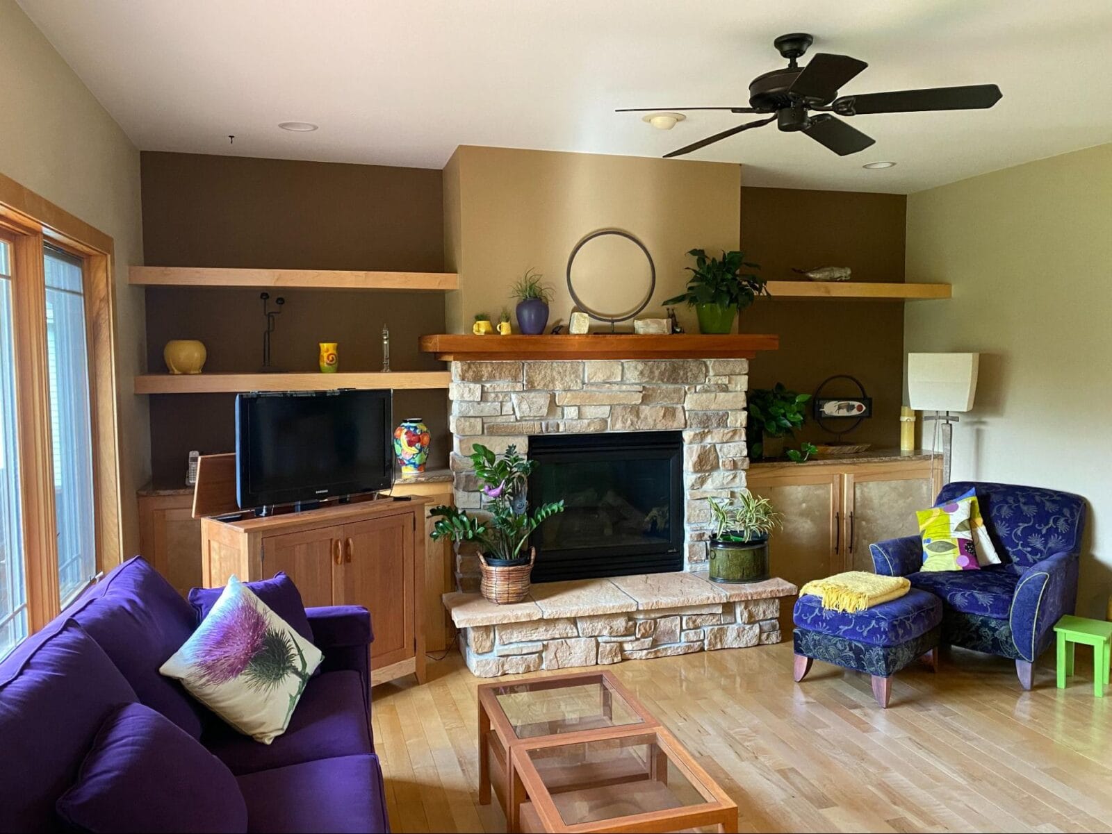 Cozy living room featuring a stone fireplace, purple couch, colorful decor, and wooden shelves with plants and art.