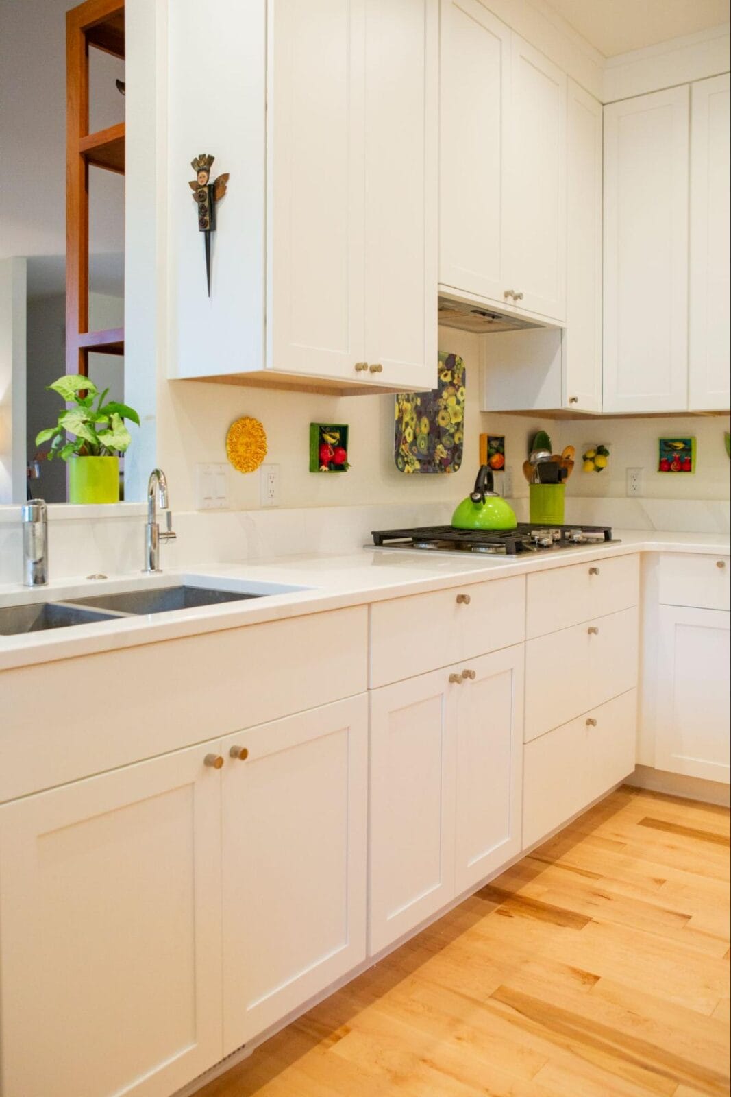 Modern kitchen with white cabinetry, stainless steel sink, green kettle, and colorful wall decor featuring plants and fruits.