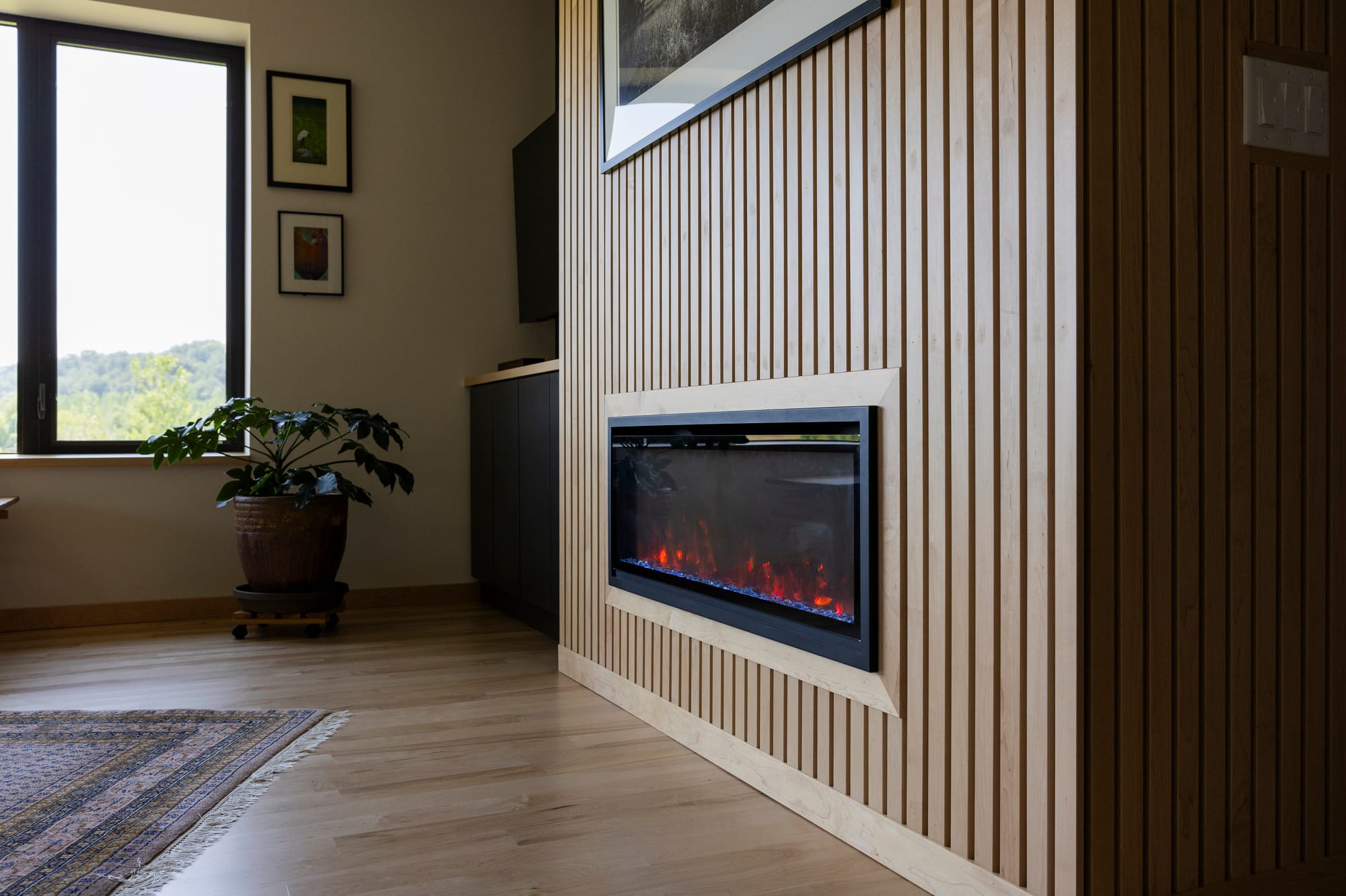Modern living room featuring a sleek electric fireplace, wooden slat wall, potted plant, and large window with scenic view.