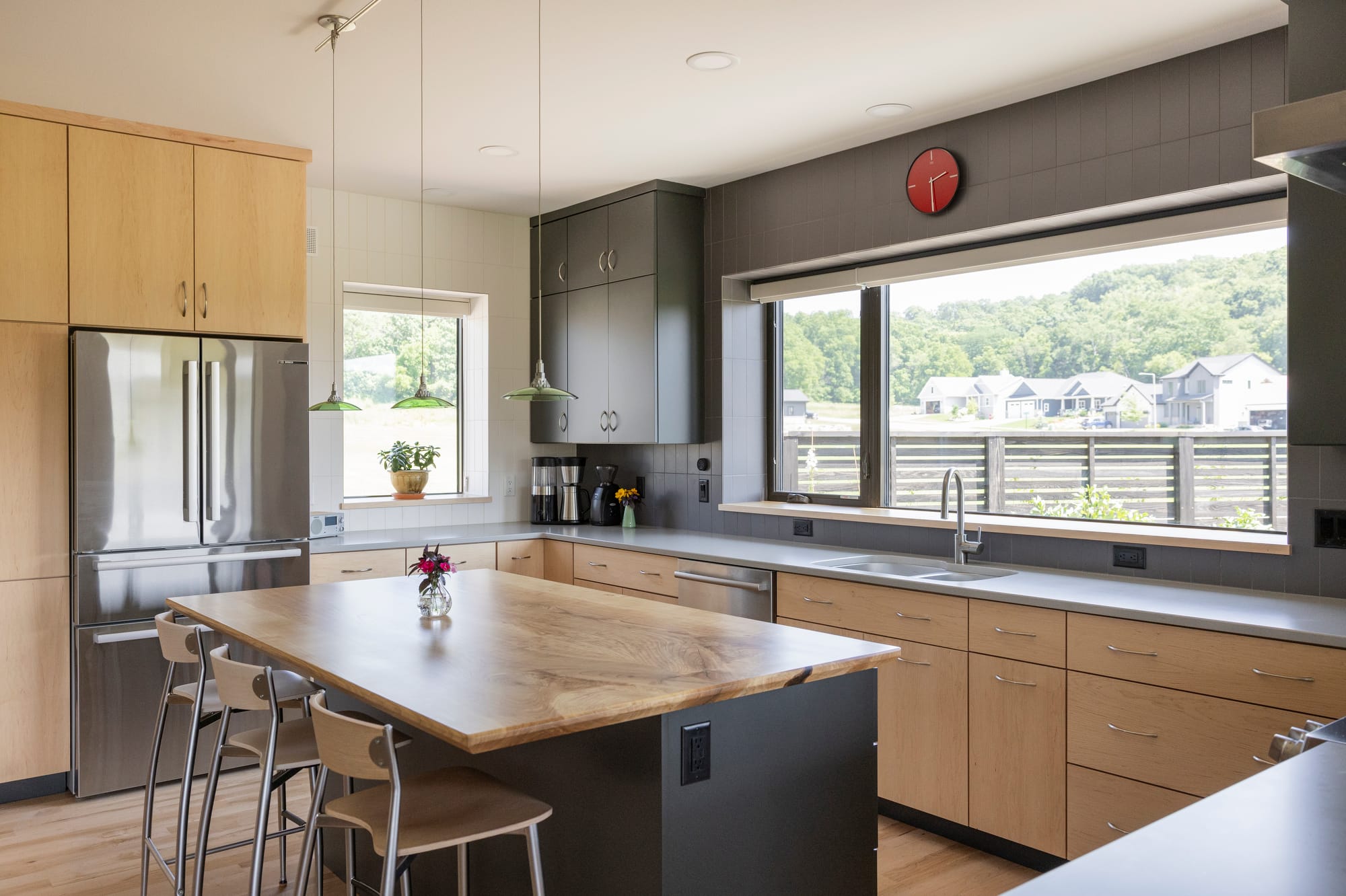 Modern kitchen featuring wooden cabinets, stainless steel appliances, and a large island with bar stools, overlooking greenery.