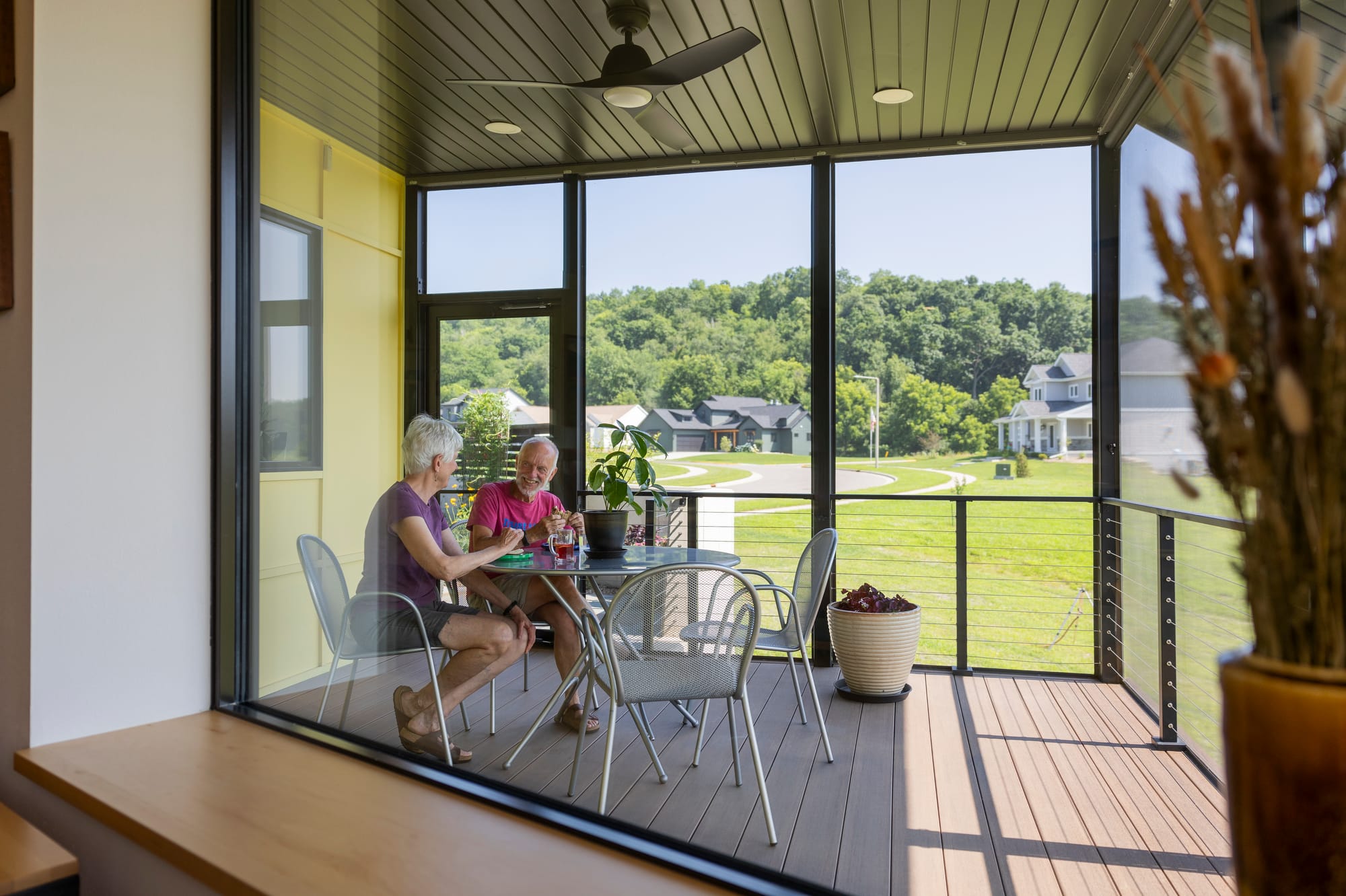 Two older adults enjoy drinks and conversation on a screened porch, surrounded by greenery and a view of nearby homes.