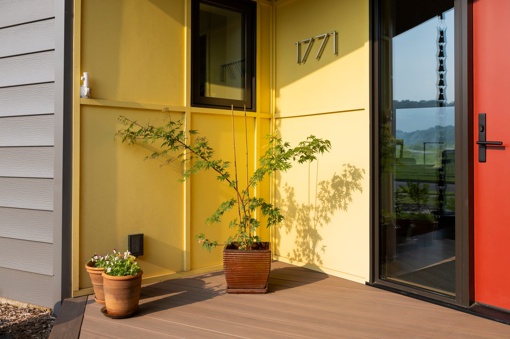 Bright yellow wall with house number 1771, potted plants, and a bold red door creating a welcoming entrance.