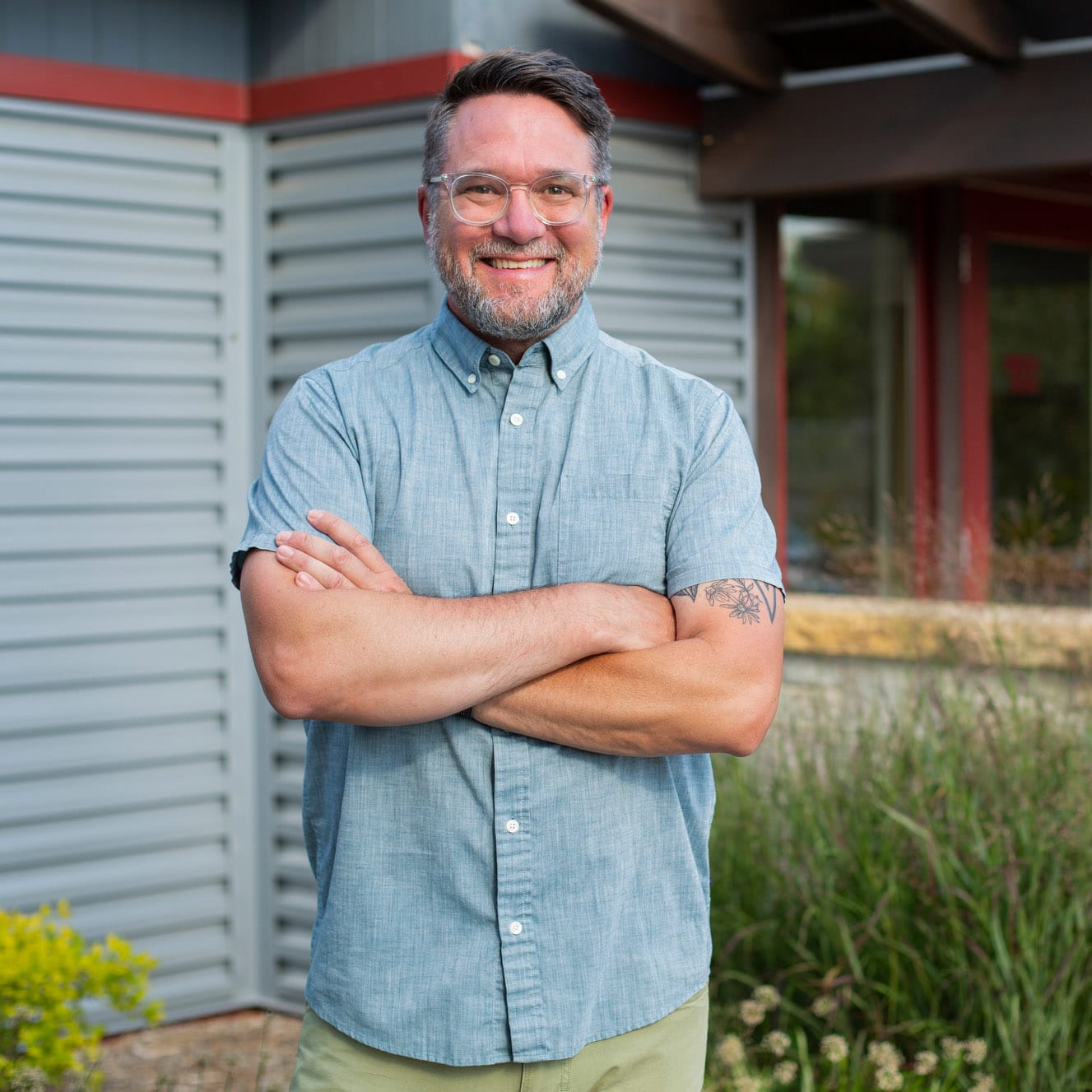 Smiling man with glasses and a beard, wearing a light blue button-up shirt, stands against building background.