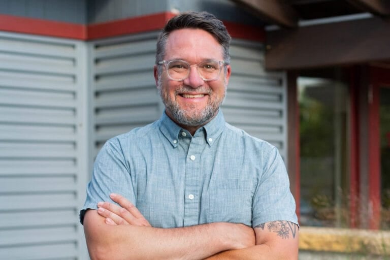 Smiling man with glasses and a beard, wearing a light blue shirt, stands with arms crossed against a modern building backdrop.