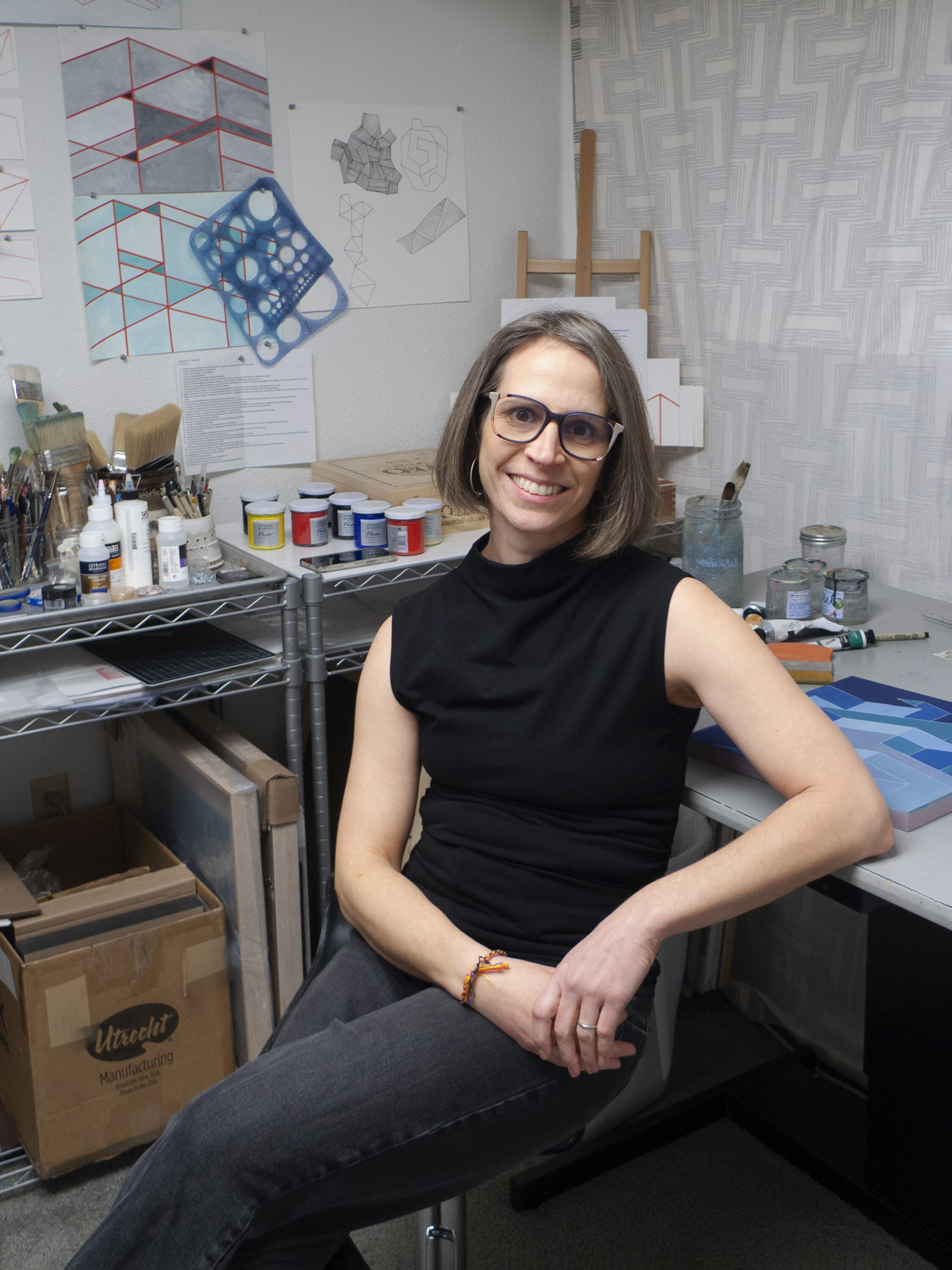 Artist in studio, surrounded by art supplies and geometric designs.