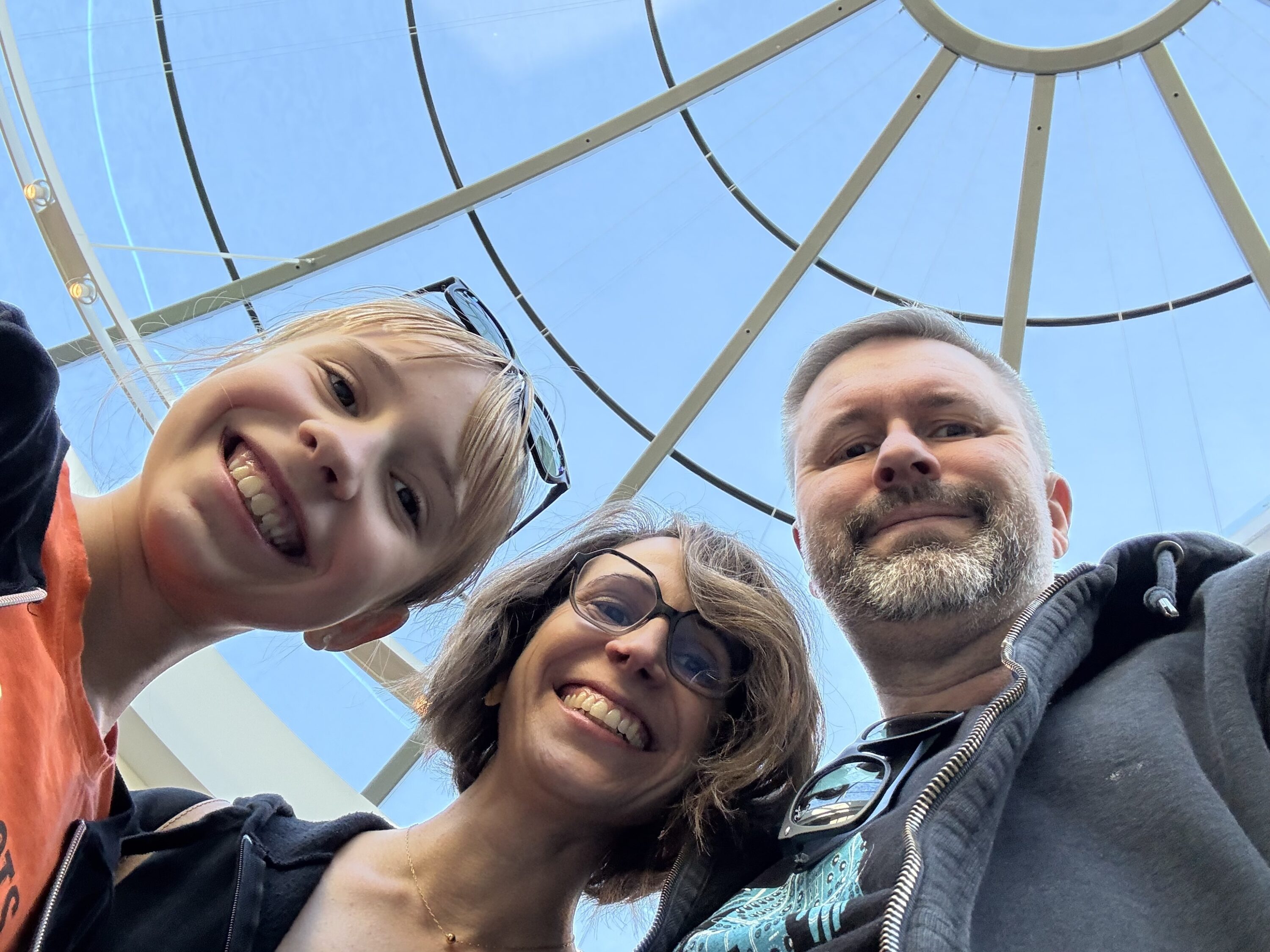 Family of three smiling up at a glass dome ceiling.