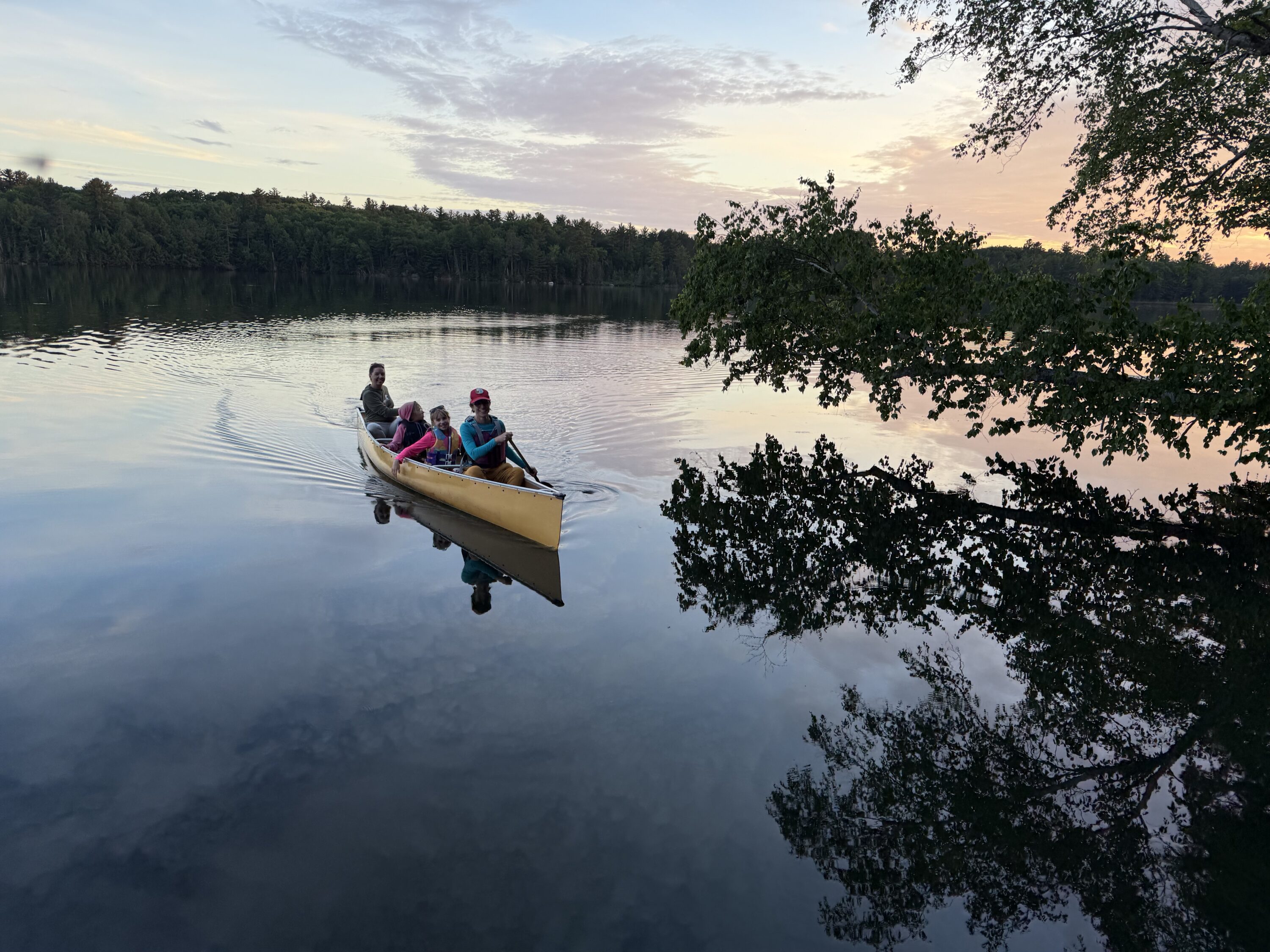 Family in a canoe on a calm lake at sunset, with trees reflected in the water.