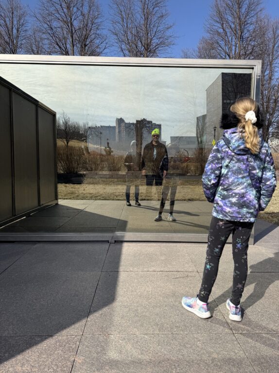 Girl in galaxy jacket looks at reflections of people in a mirrored wall.