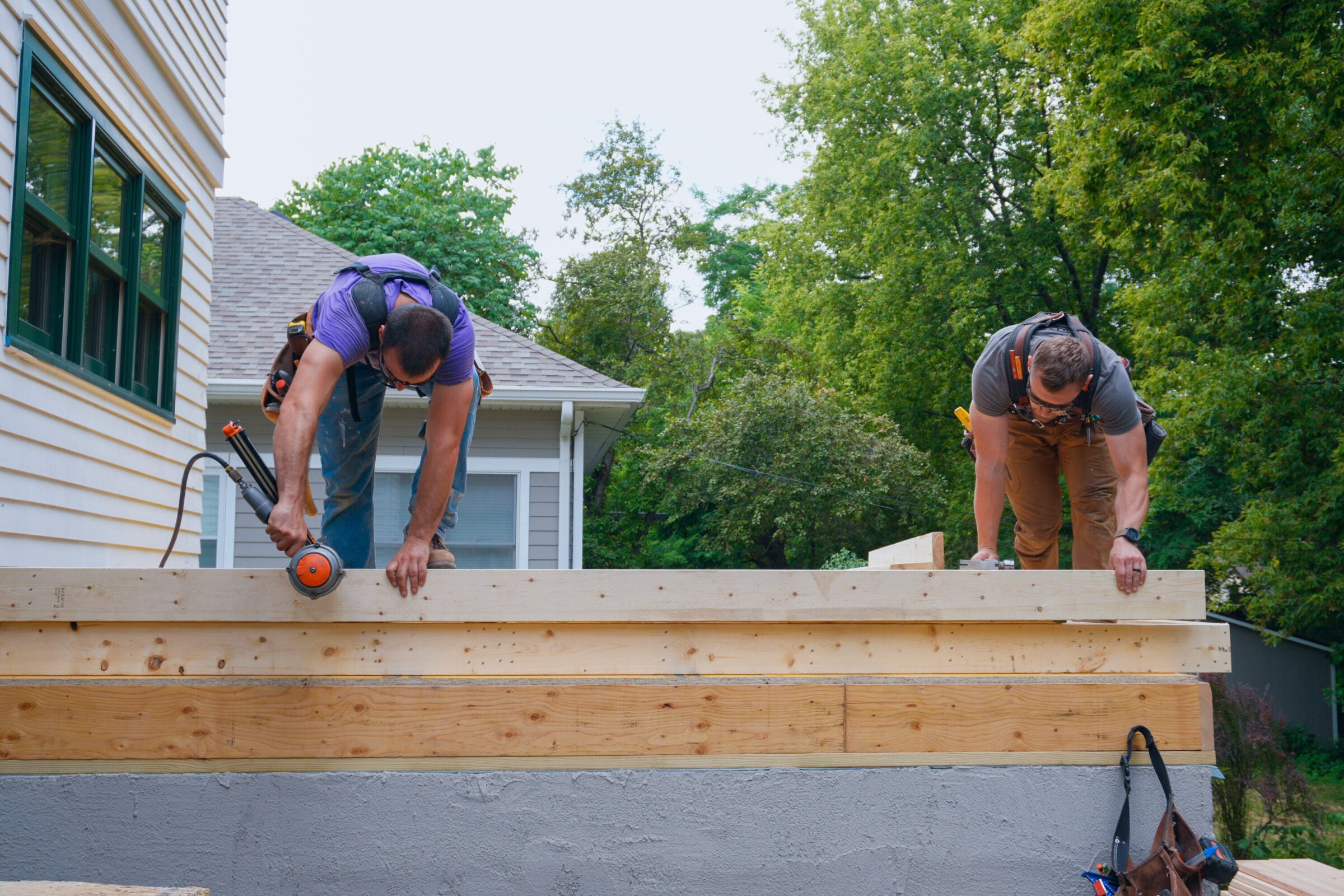 Two construction workers build a wooden deck, one using a nail gun.