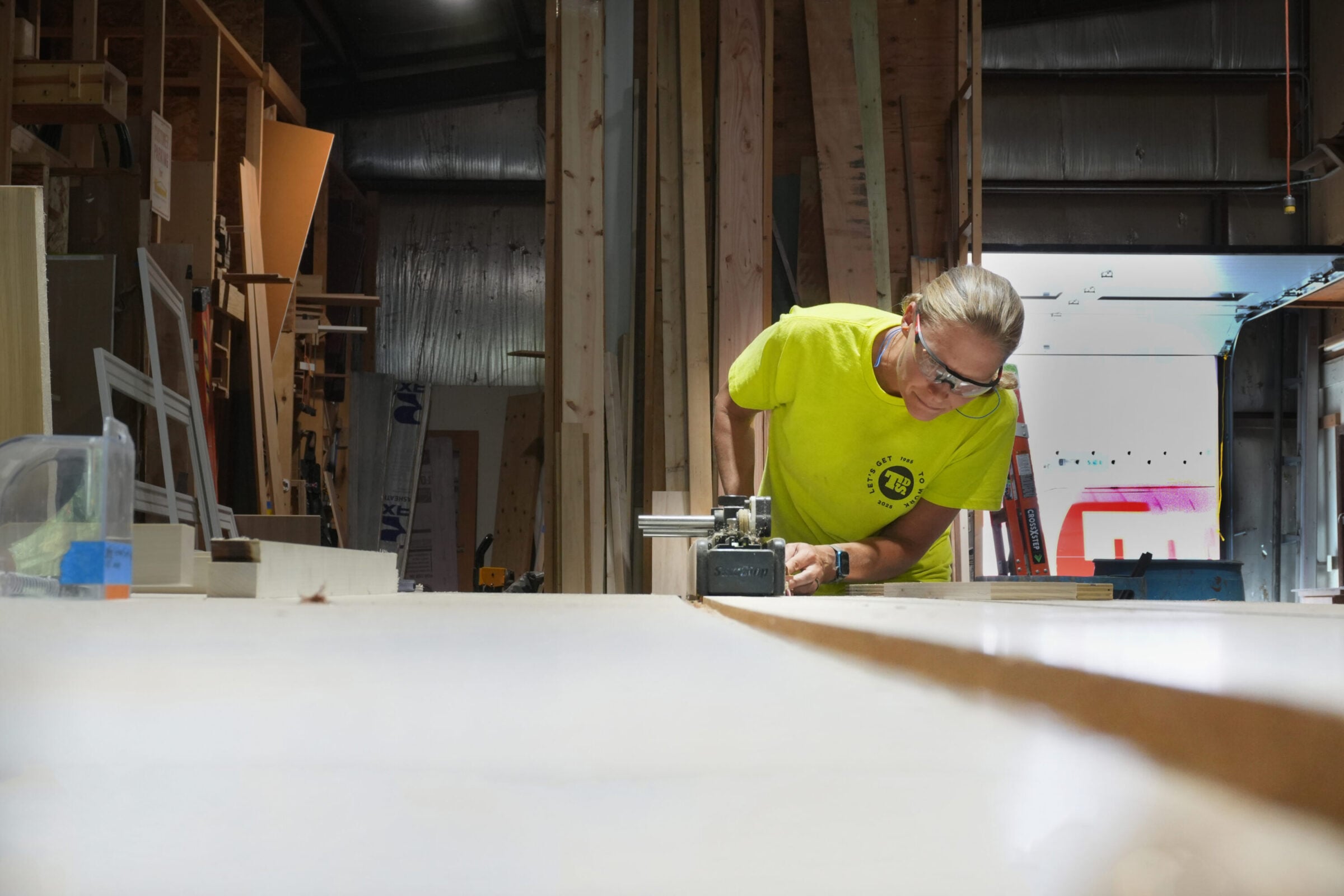 Woodworker in yellow shirt and safety glasses uses a router on a large piece of wood.